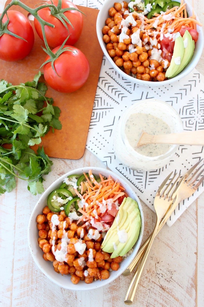 bowls filled with chickpeas tossed in buffalo sauce and sliced vegetables with two forks and a jar of ranch dressing on the side