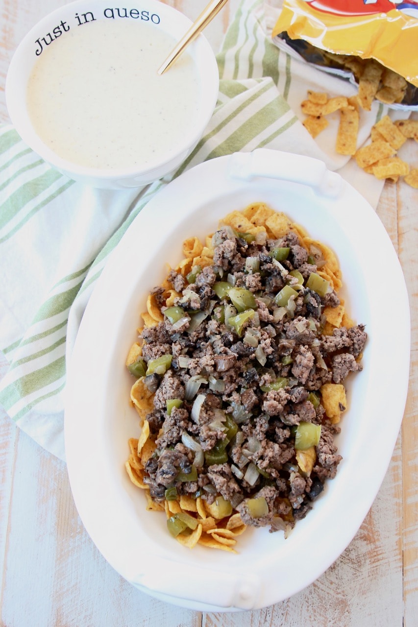 Frito pie topped with ground beef with a bowl of provolone cheese sauce and bag of fritos