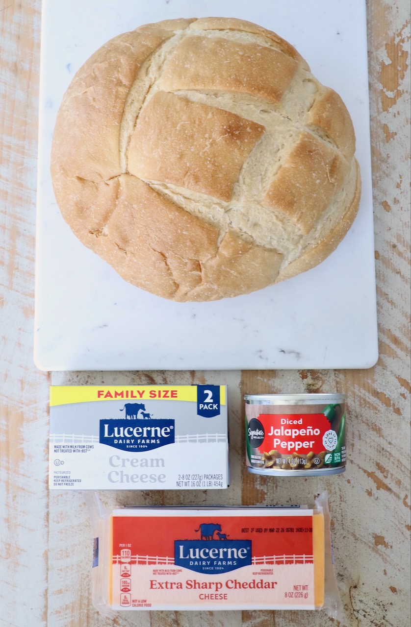 Round loaf of bread on a marble board, with Lucerne cream cheese, extra sharp cheddar, and diced jalapeño peppers laid out on a rustic wooden table.