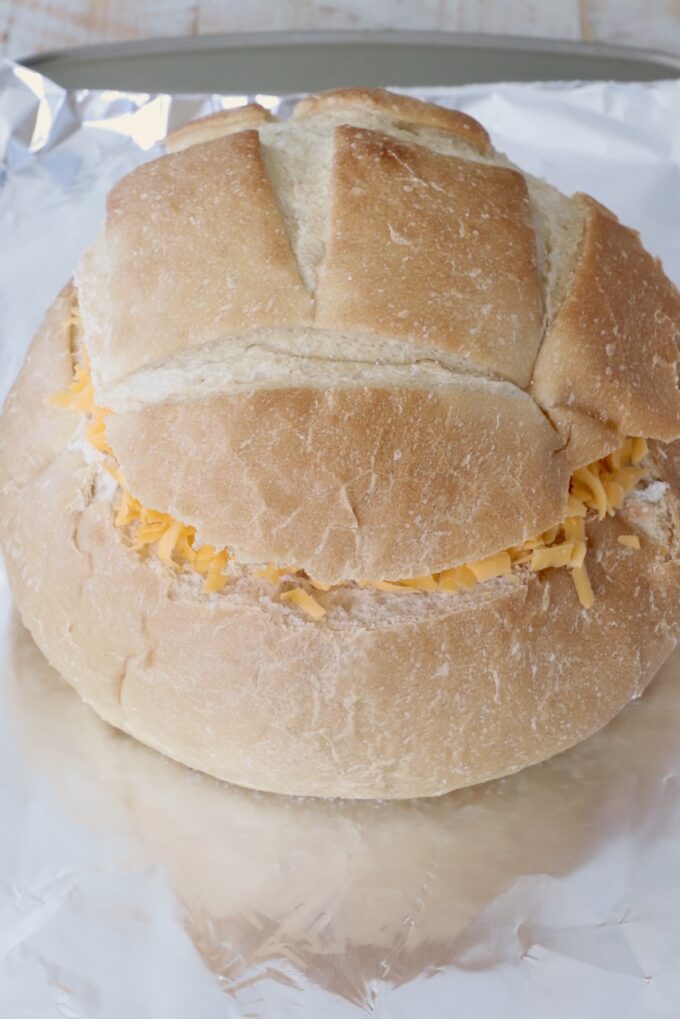 A round bread loaf with its top sliced off, filled with shredded cheese, rests on a sheet of aluminum foil on a baking sheet.