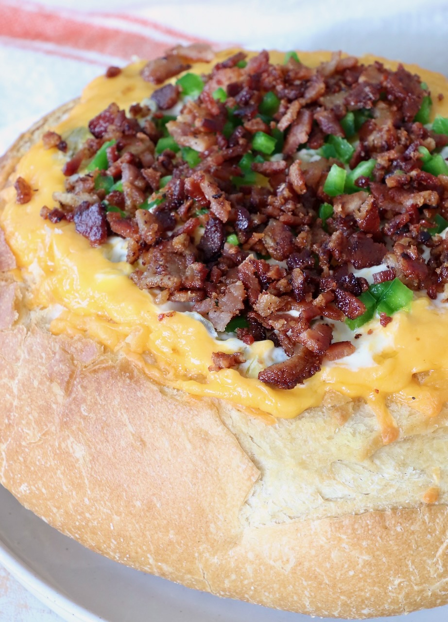 Round bread loaf hollowed and filled with melted cheese, topped with crispy bacon bits and chopped green peppers