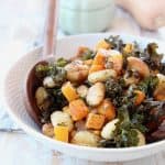Baked gnocchi, butternut squash and kale in bowl with wooden serving spoons, whole butternut squash and green baking dish in back