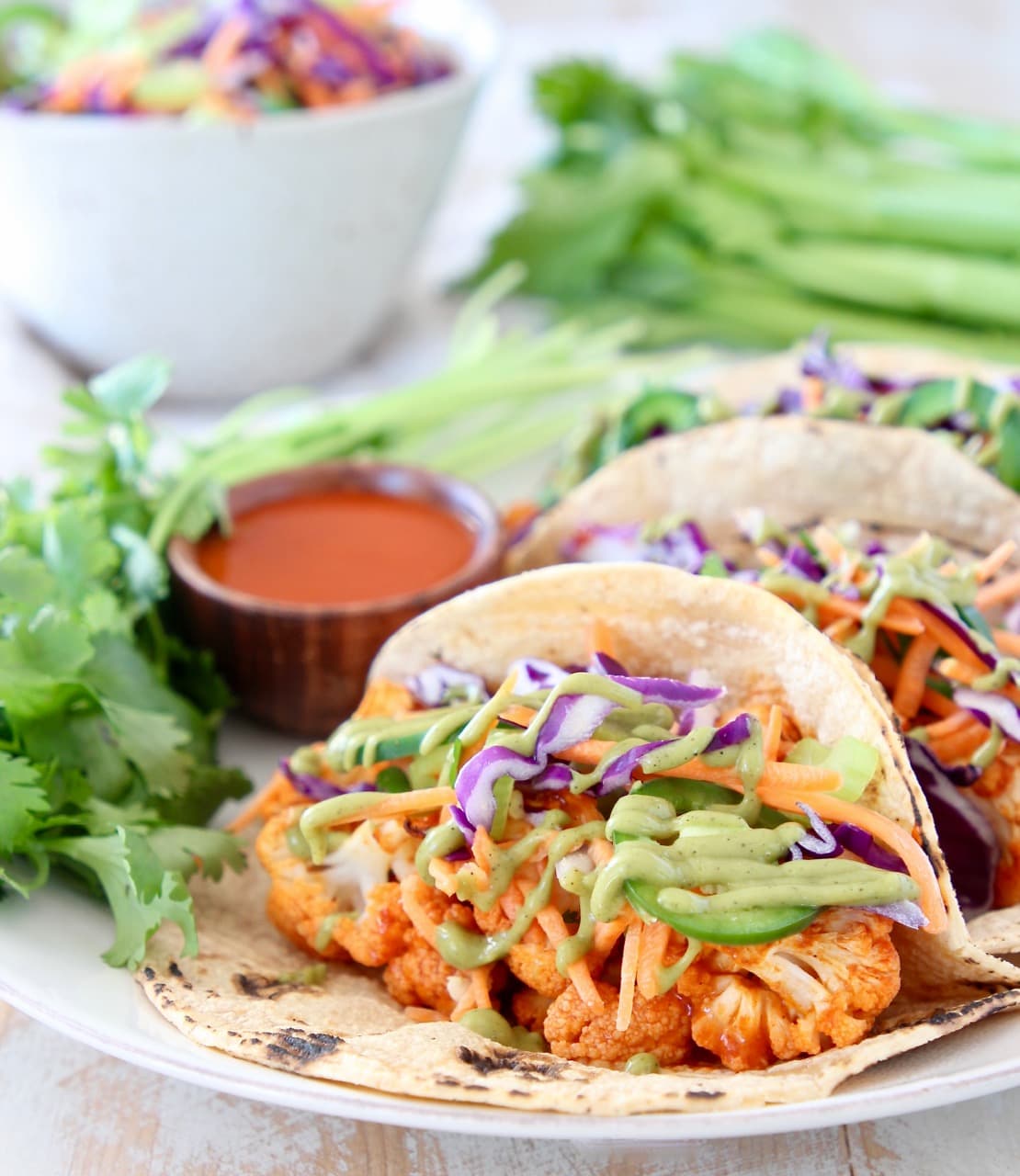 Buffalo Cauliflower Tacos in Corn Tortillas with Jalapeno Slaw and Avocado Sauce on plate with small bowl of buffalo sauce and fresh cilantro