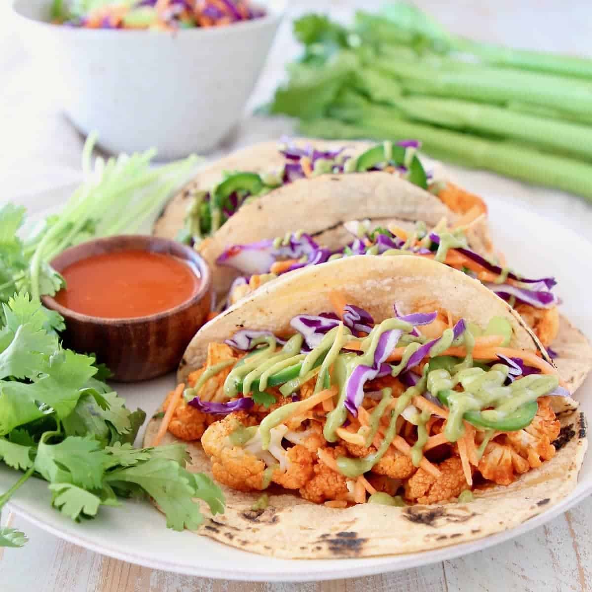 Buffalo cauliflower tacos on corn tortillas with jalapeno carrot slaw and fresh cilantro on plate with small bowl of buffalo sauce