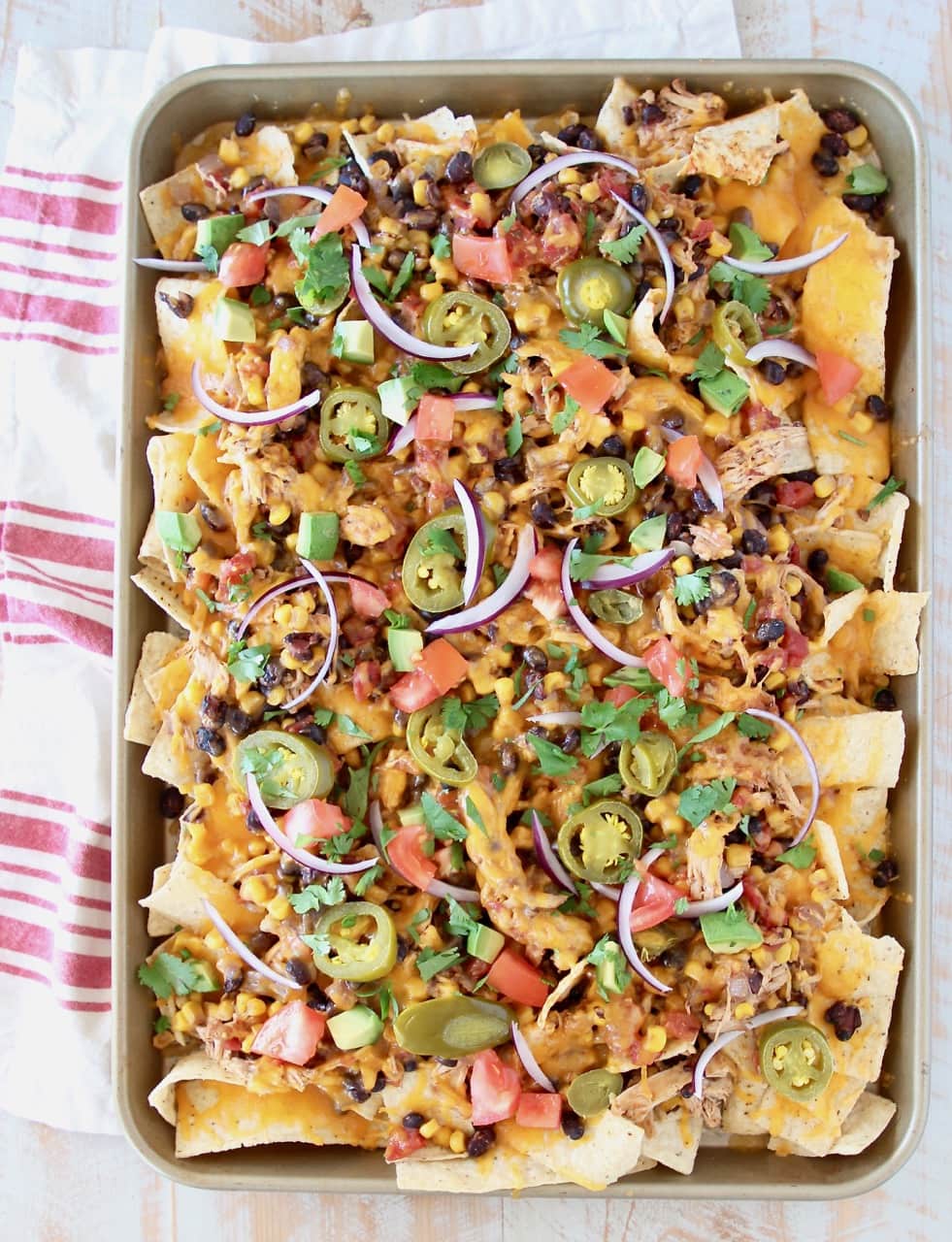 Sheet pan of chicken nachos topped with cheese, tomatoes, avocado, onions and jalapenos on red and white striped towel