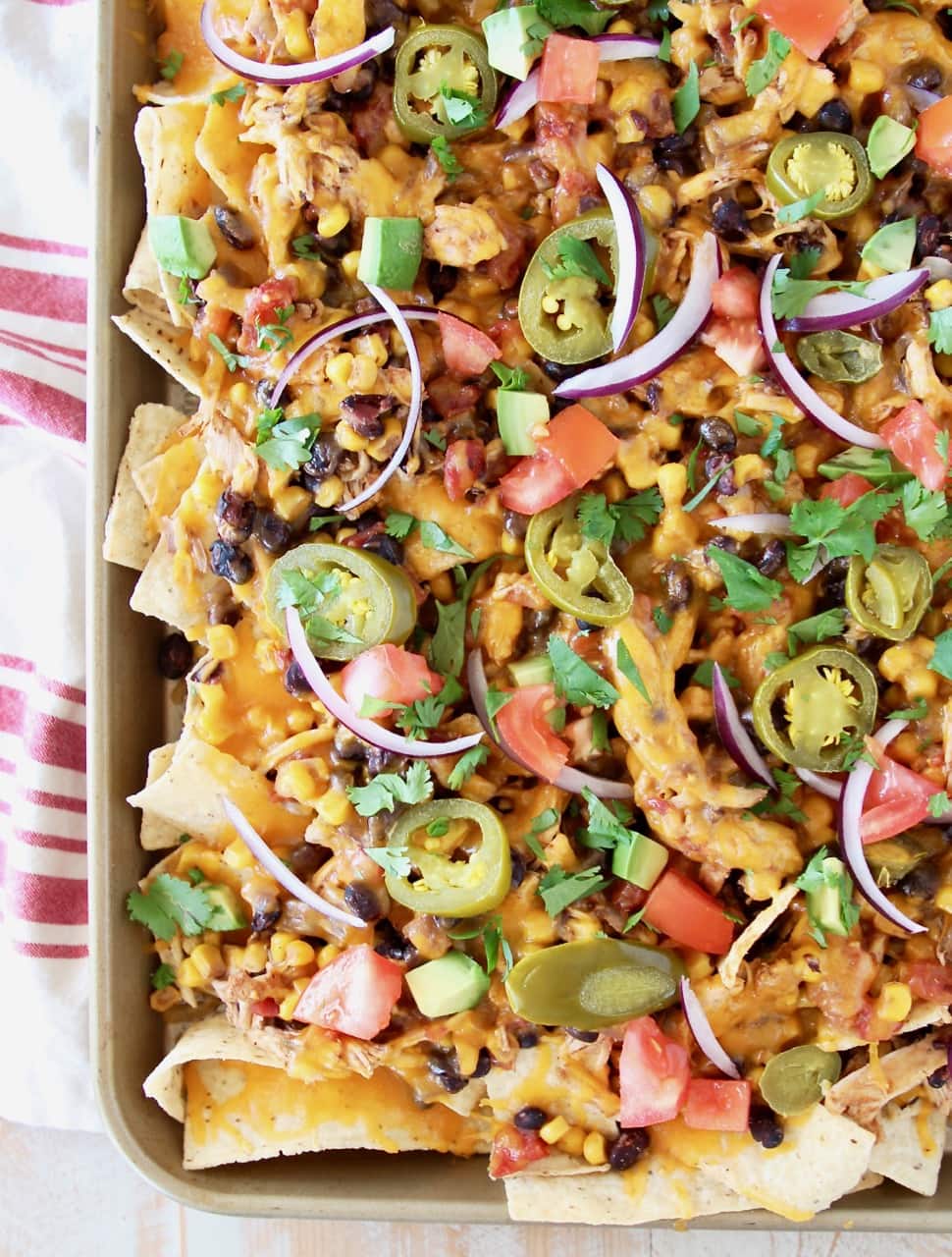 Chicken Nachos on sheet pan topped with jalapenos, red onions, tomatoes and cheese, on red and white striped towel