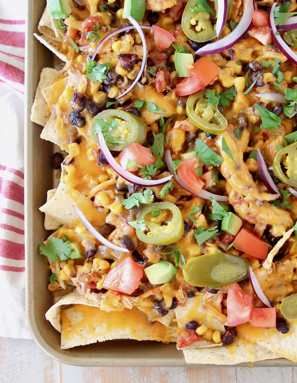 Chicken nachos on sheet pan with melted cheddar cheese, diced tomatoes, red onions and cilantro on red and white striped towel