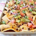 Chicken nachos on baking sheet with jalapenos, tomatoes, cheese, black beans and red onions, sitting on red and white striped towel