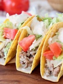 Ground beef tacos in crispy shells with diced tomatoes and shredded lettuce on wood cutting board with whole tomatoes in back