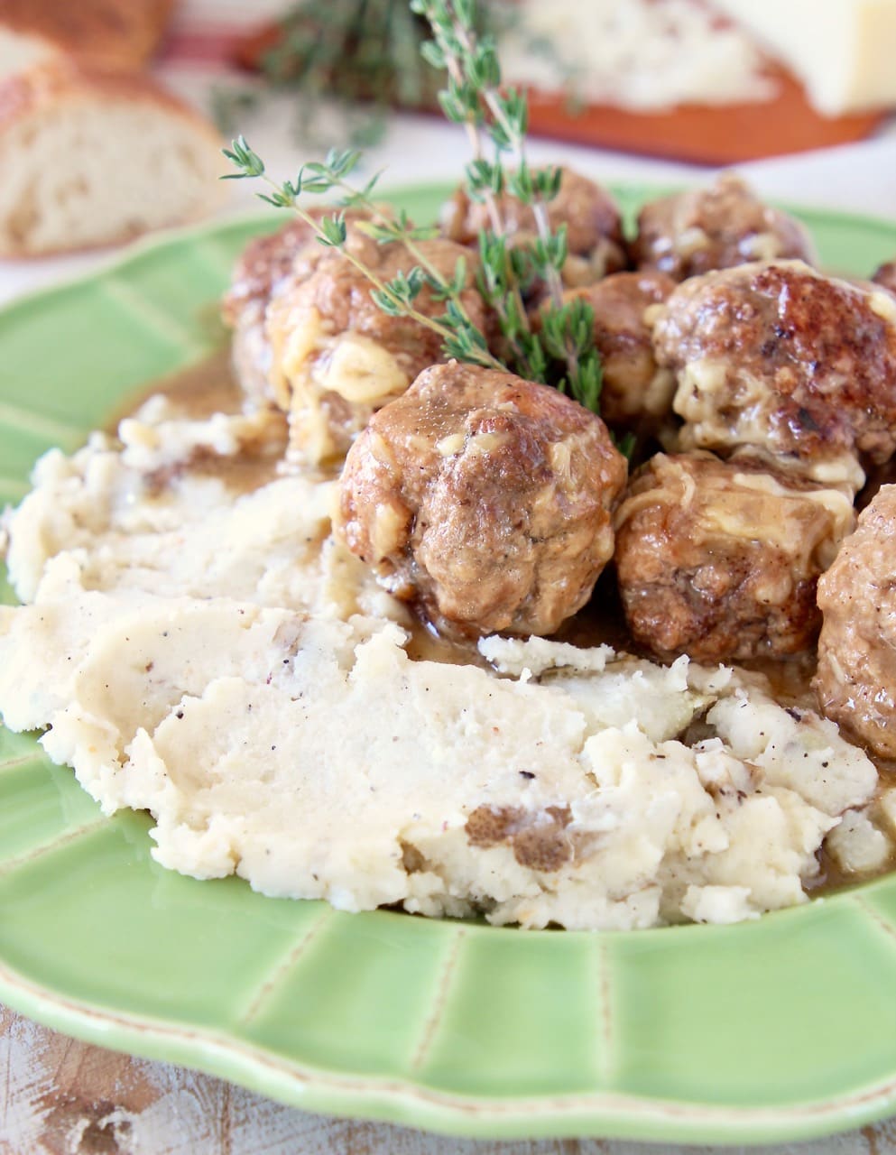 Green plate with mashed potatoes and meatballs, a fresh sprig of thyme and sliced baguette in the background