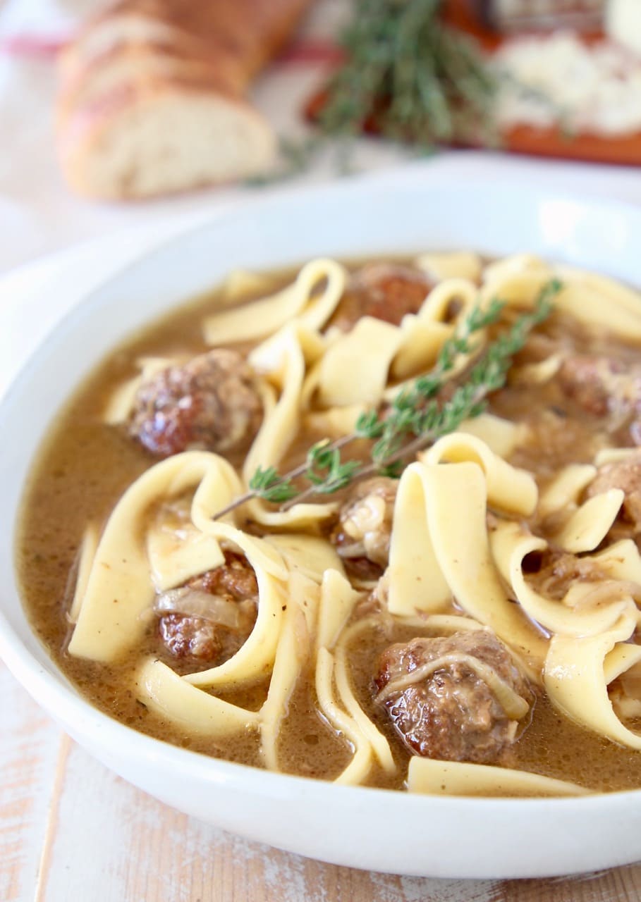 Meatballs and egg noodles in french onion soup in large white bowl with sprigs of fresh thyme and a sliced baguette in the background