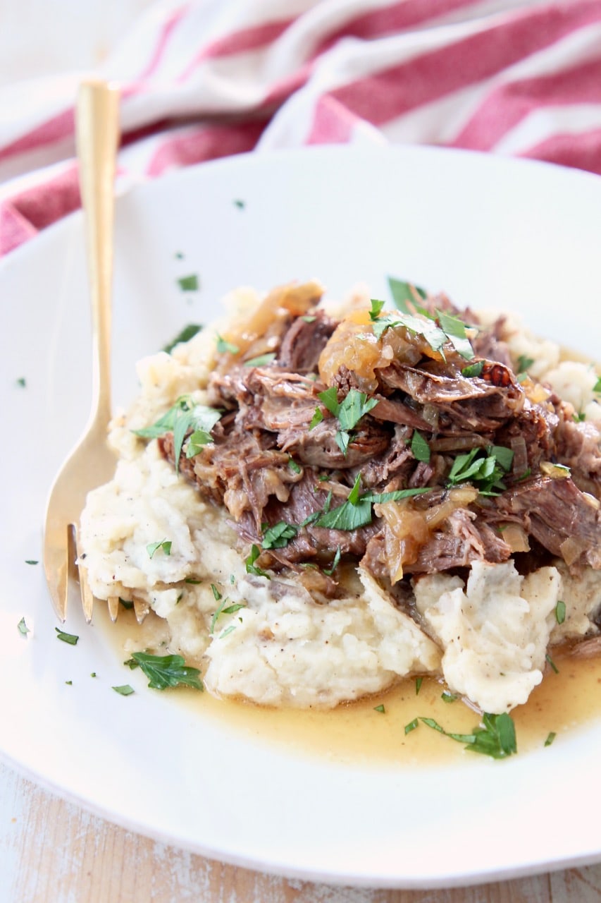 Shredded pot roast with onions on top of mashed potatoes on white plate with gold fork and red and white striped towel