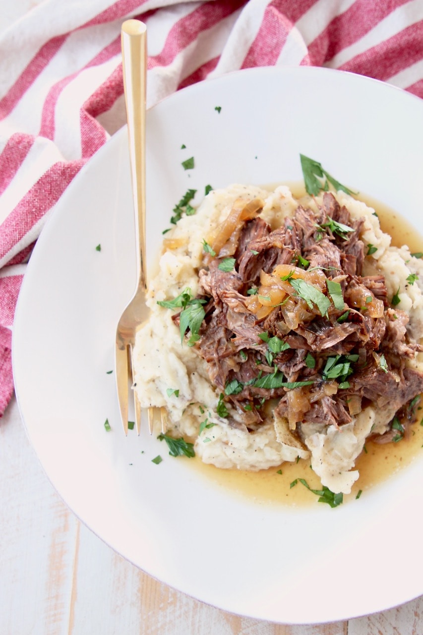 Shredded french onion pot roast on top of mashed potatoes on a plate, topped with fresh chopped parsley with a gold fork and red and white towel