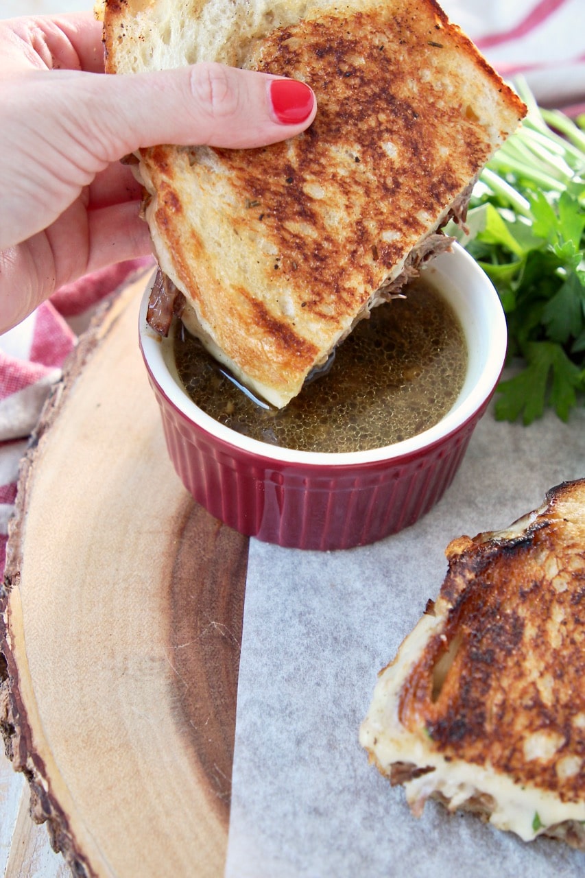 Grilled cheese pot roast sandwich held in hand, being dipped into a red ramekin of au jus