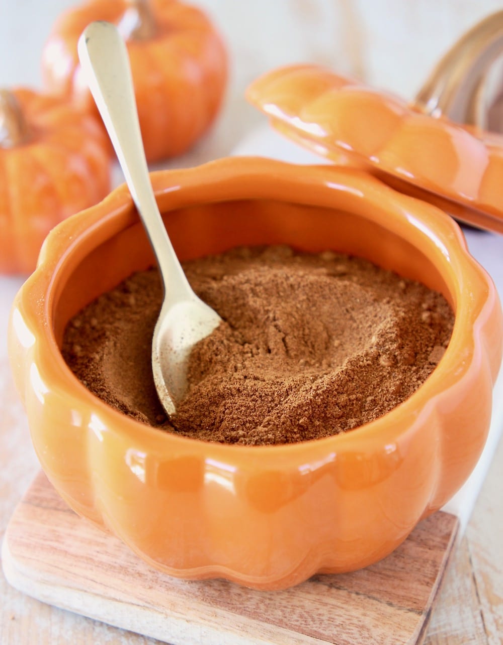 Homemade pumpkin pie spice in an orange pumpkin bowl with a small gold spoon and two small ceramic pumpkins