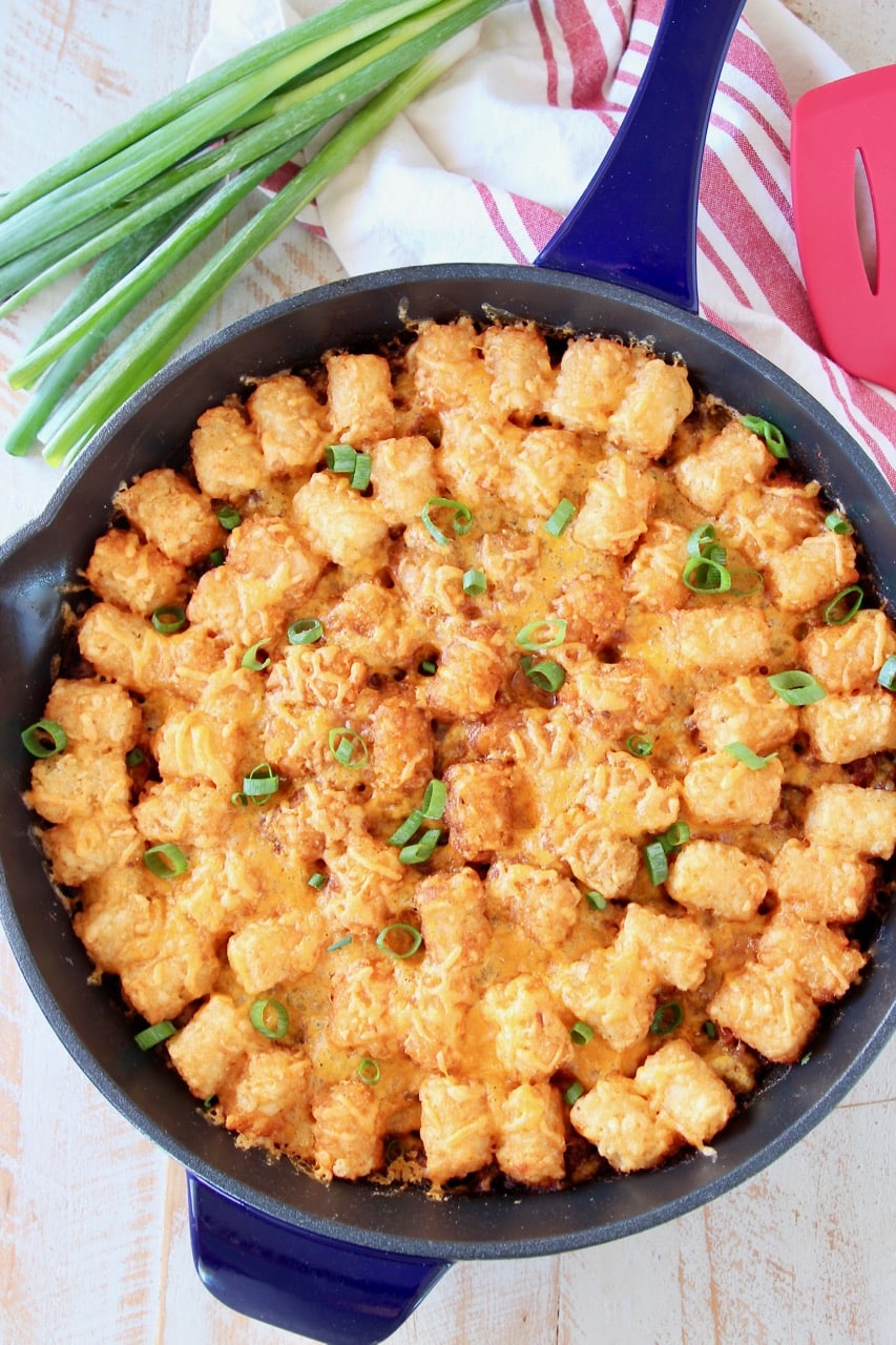 The best Tater Tot Casserole with ground beef and cheese in skillet with green onions and red striped towel