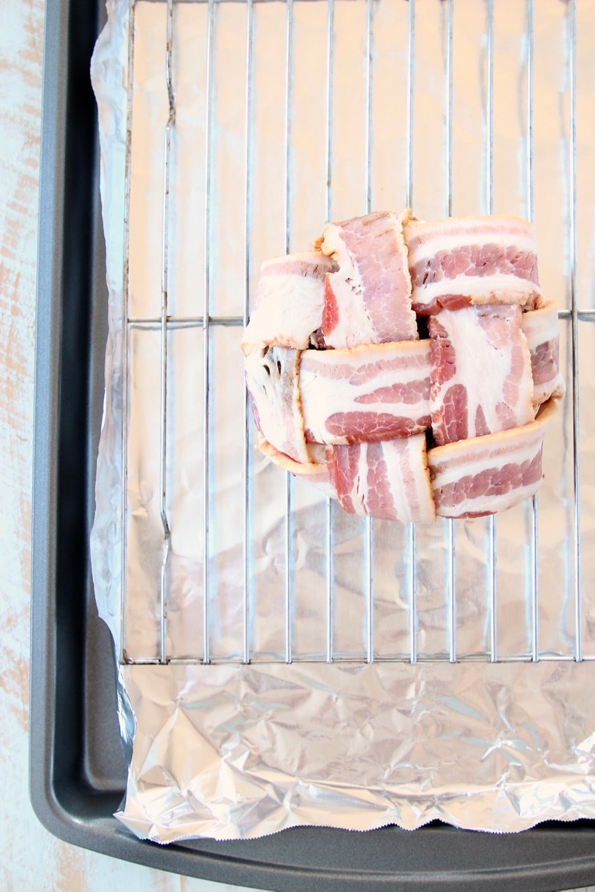 Bacon weave wrapped around a round of brie cheese on a wire rack on top of a foil lined baking sheet