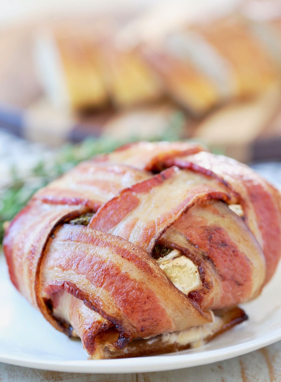 Baked bacon wrapped brie with bacon weave on plate with fresh herbs, cutting board with sliced baguette in background
