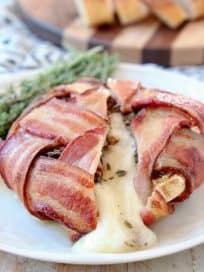 Bacon Wrapped Brie with melted brie spilling out onto plate with fresh herbs on plate and sliced baguette on wood cutting board in background
