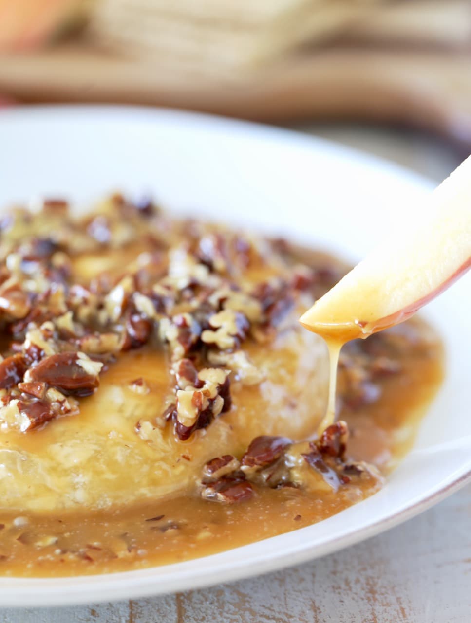 Apple slice dipped into salted caramel poured over baked brie with pecans on white plate