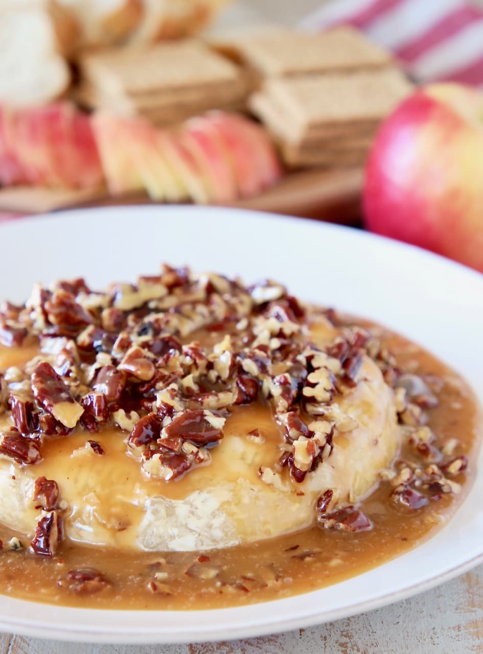 Easy baked brie recipe topped with salted caramel sauce and pecans on a white plate with a black cheese spreader, with apples in the background 