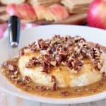 Baked brie round topped with salted caramel sauce and pecans on white plate with black cheese spreader and wooden tray of sliced apples and graham crackers in background