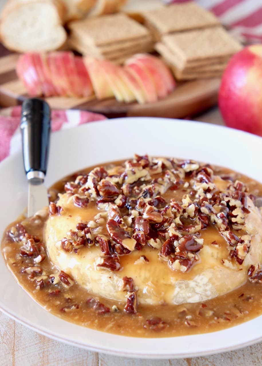 Salted Caramel and pecans on top of baked brie round on plate with cheese spreader and apples, graham crackers and sliced baguette in the background