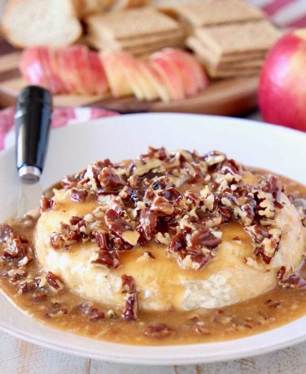 Salted Caramel and pecans on top of baked brie round on plate with cheese spreader and apples, graham crackers and sliced baguette in the background