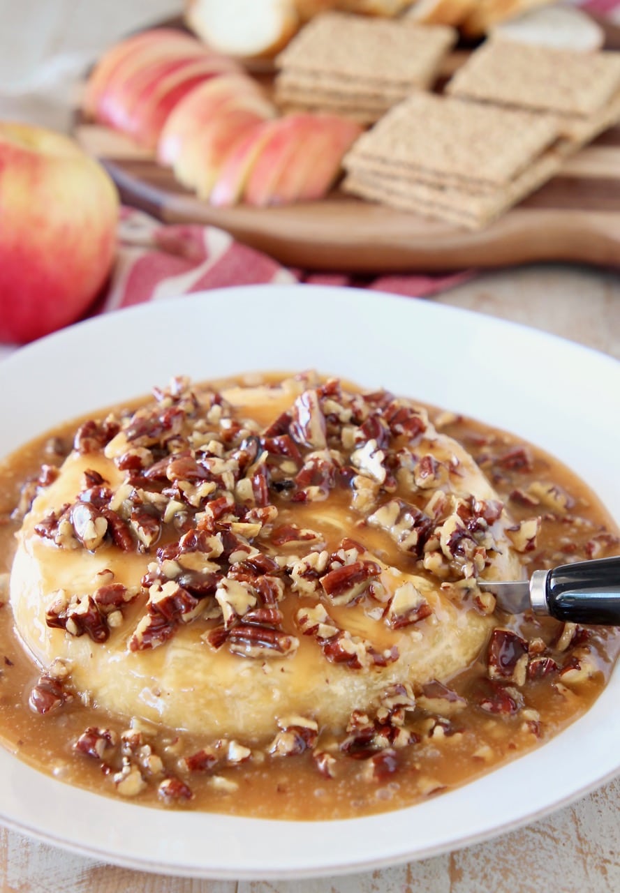 Salted caramel and pecans poured over a baked brie round on a baking sheet with a black cheese spreader slicing into the baked brie