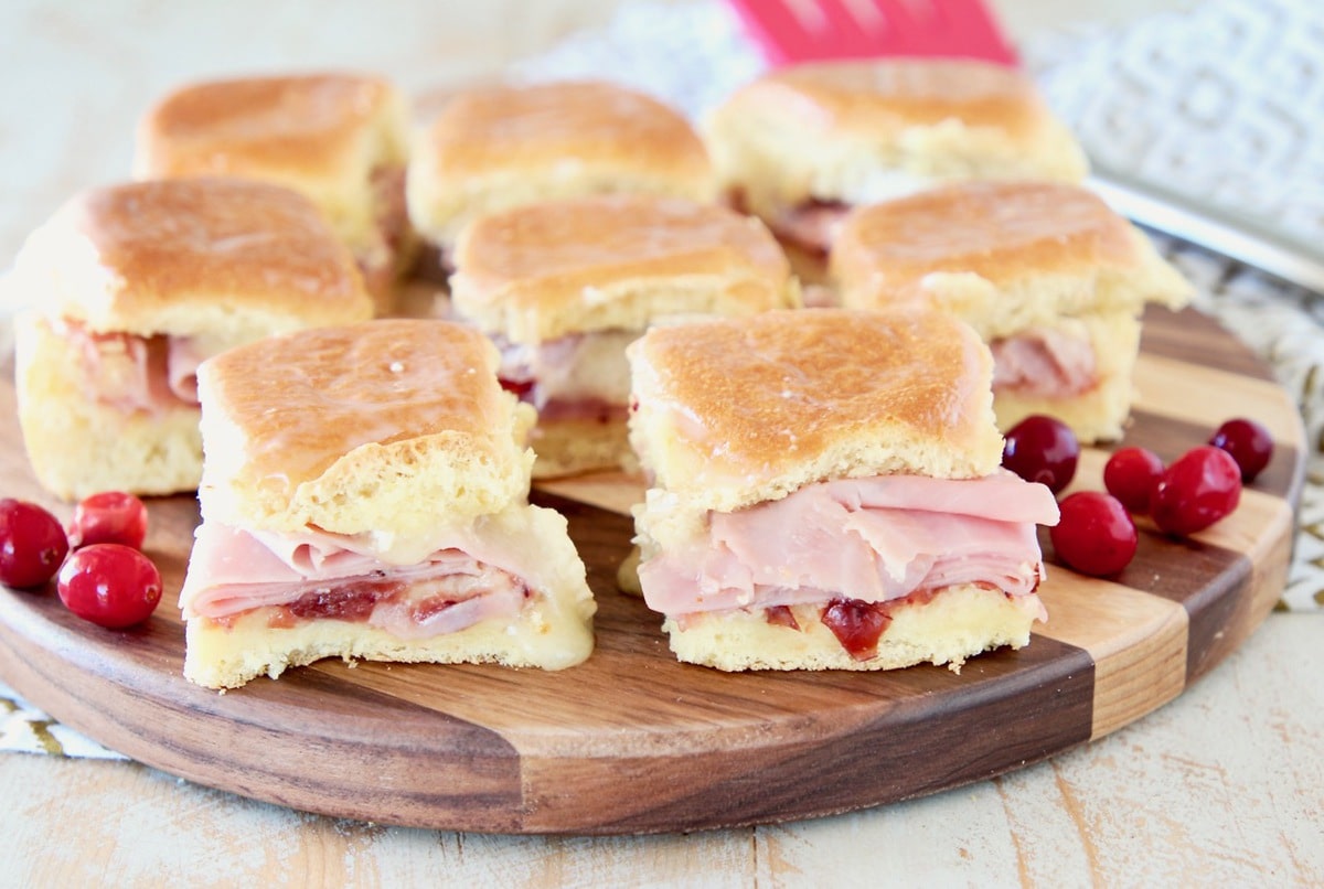 Ham sliders with cranberry sauce and brie cheese on hawaiian rolls, sitting on a wood cutting board with red spatula in background