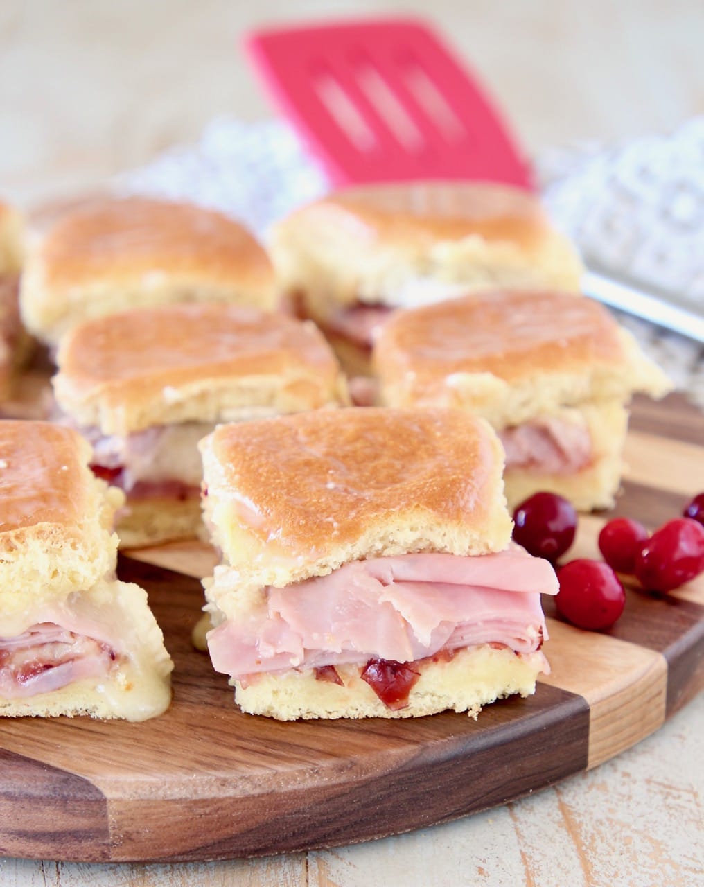 Ham sliders on hawaiian rolls with cranberry sauce, on wood cutting board with fresh cranberries on the side and red spatula in the background