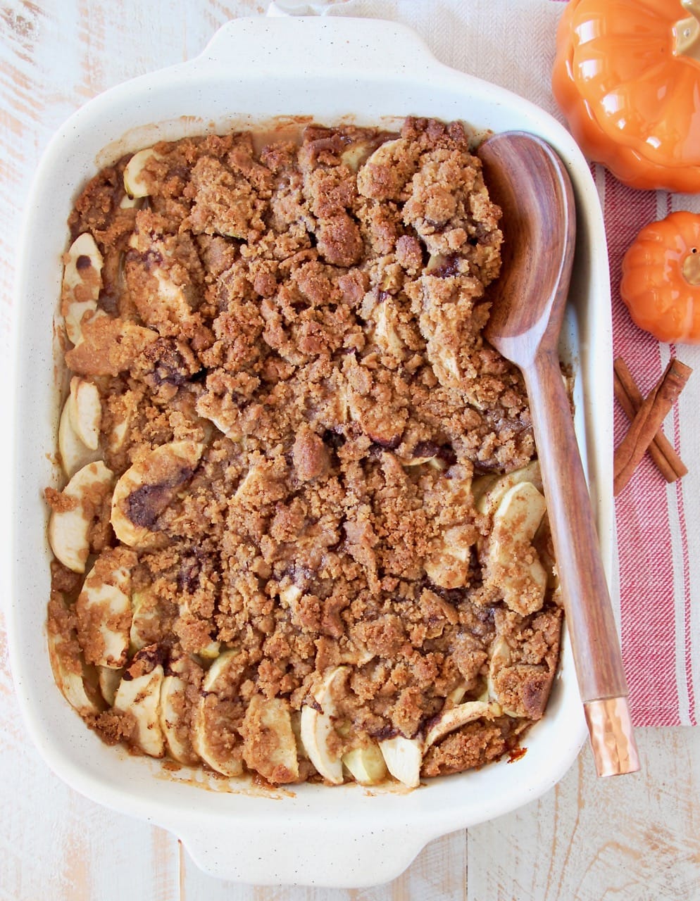Overhead shot of apple crumble in a white casserole baking dish with a wooden spoon, sitting next to small orange pumpkins and cinnamon sticks on a red and white striped towel