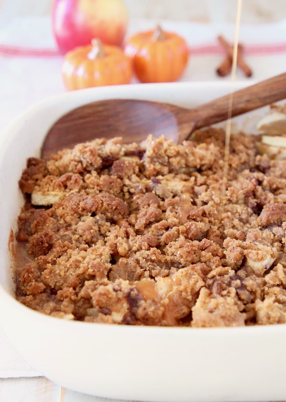 Pumpkin spice apple crumble in casserole dish with wooden spoon and drizzle of caramel syrup, with small ceramic pumpkins in background