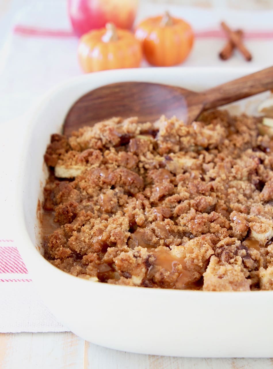 Apple crumble in baking dish with wooden spoon on white and red striped towel with small orange pumpkins in background