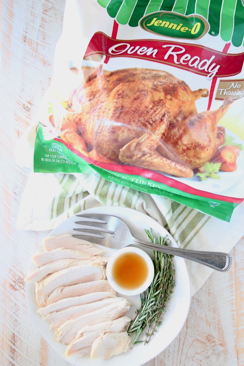 Plate of sliced roasted turkey with large fork, bowl of maple apple reduction and fresh rosemary springs, next to Jennie O Whole Oven Ready Turkey in bag