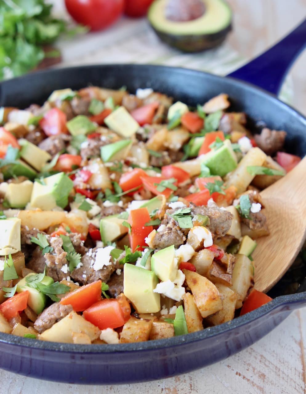 Breakfast hash in blue cast iron skillet with diced avocado, tomatoes, sausage and potatoes, with wooden spoon in skillet and fresh cilantro in background