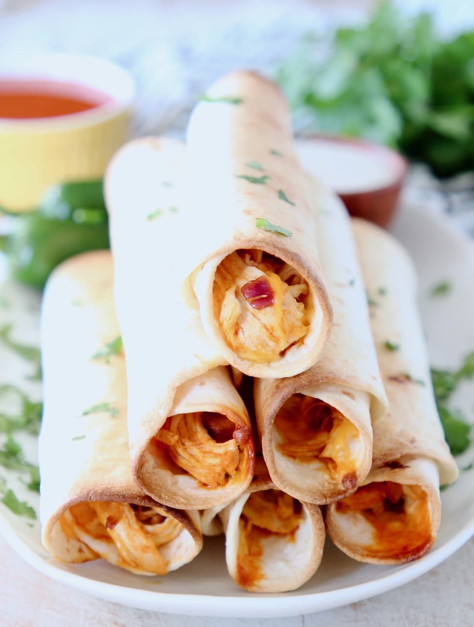 Buffalo chicken taquitos stacked up on plate with fresh cilantro and yellow ramekin of buffalo sauce in the background