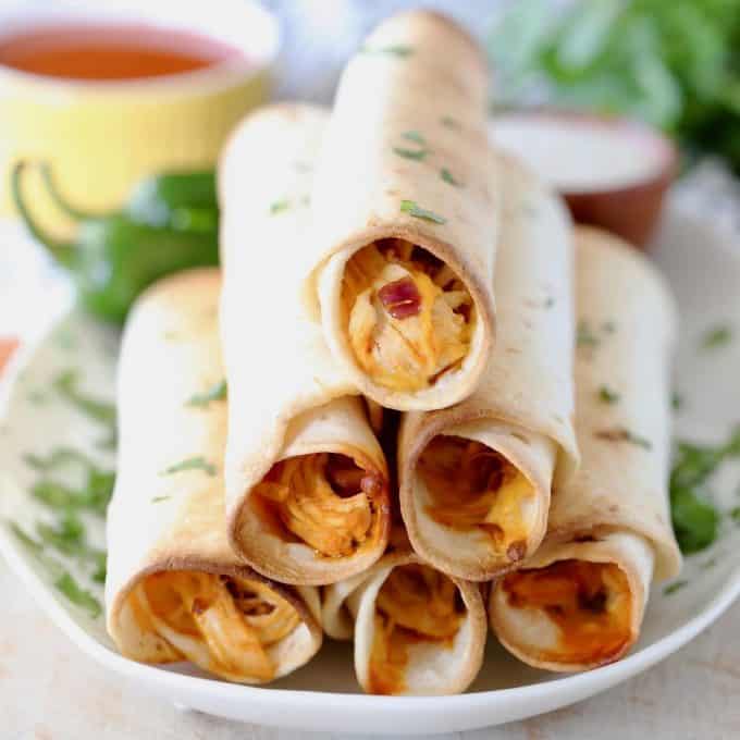 Buffalo chicken taquitos stacked on white plate with fresh cilantro garnish and jalapenos on plate in background