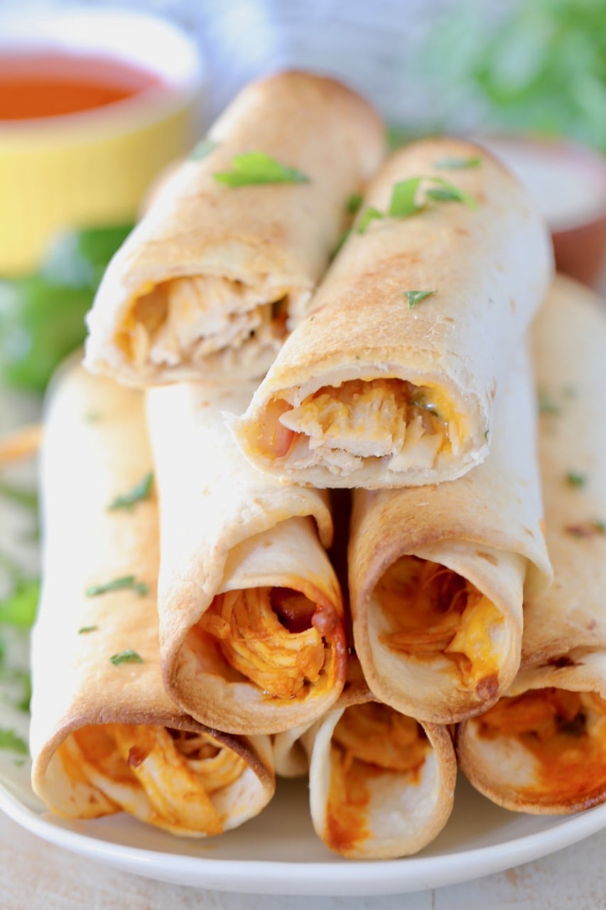 Baked buffalo chicken taquitos stacked up on a plate with the top taquito cut in half, garnished with fresh cilantro