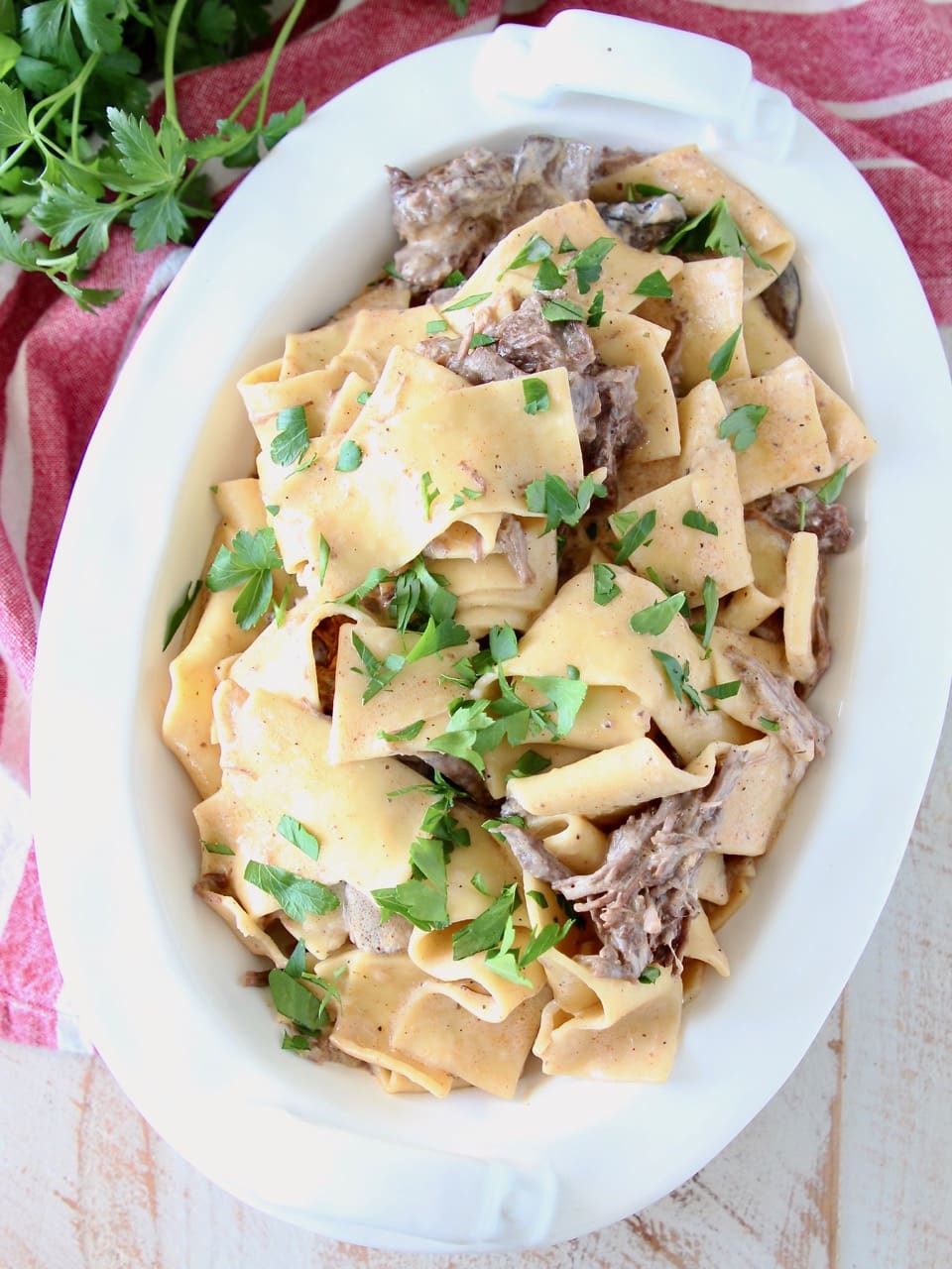 Instant pot beef stroganoff in a large white oval serving dish with wide egg noodles and shredded beef, topped with fresh parsley, sitting on a red and white striped towel