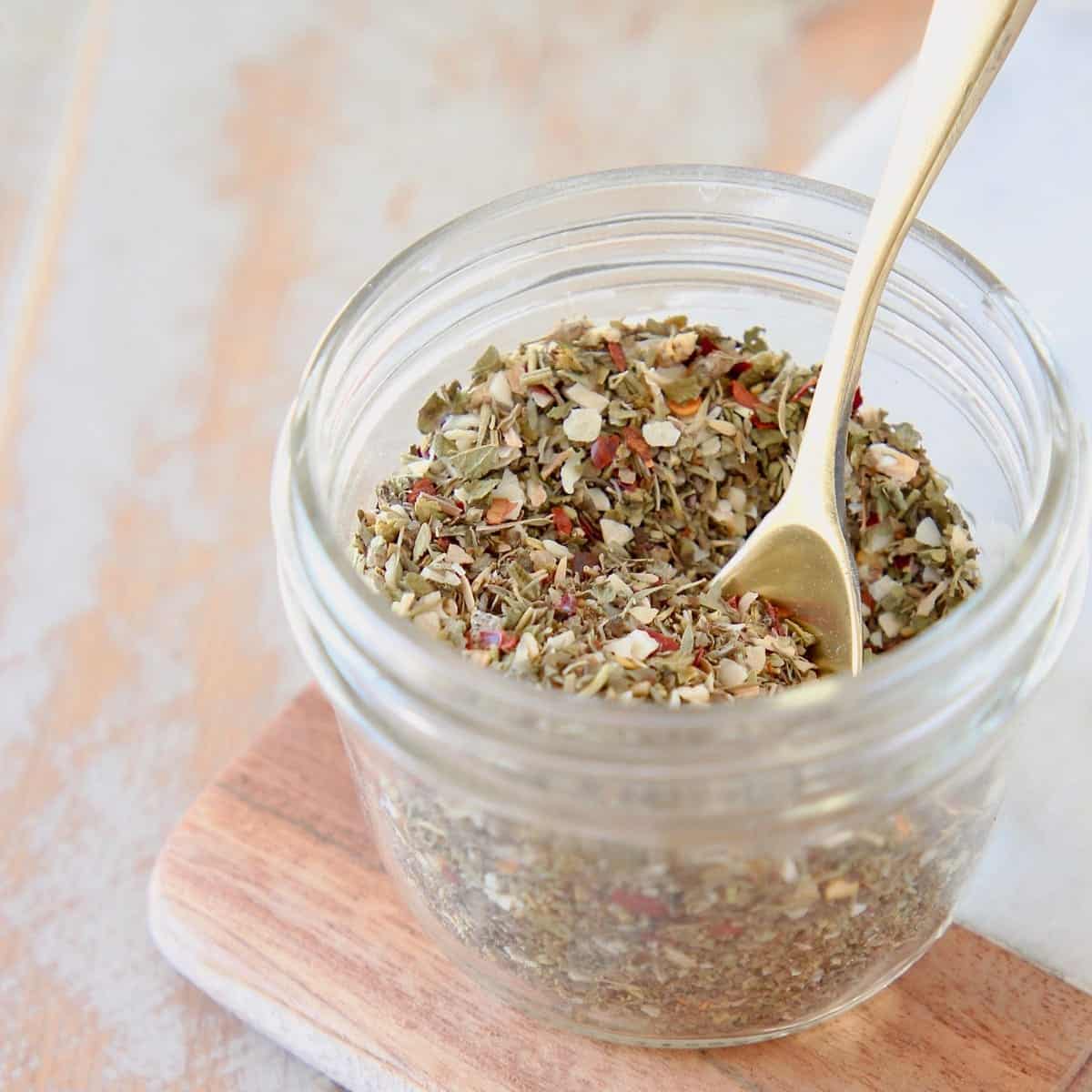 Mason jar of homemade Italian seasoning on a wood and marble cutting board with a small gold spoon in the jar