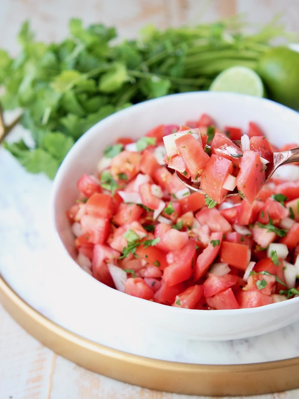 Pico de gallo in a white bowl on a white marble serving tray with gold rim, fresh cilantro and limes in background