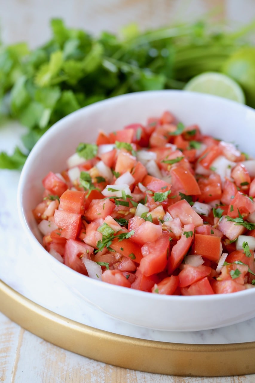 Pico de gallo in white bowl on marble serving tray, with fresh cilantro and limes in the background