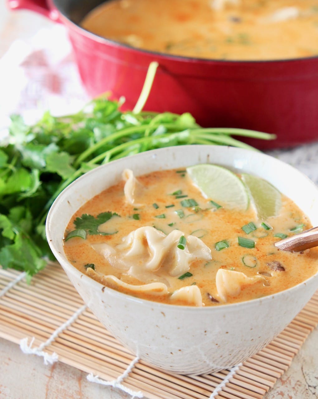 Thai coconut soup with potstickers in bowl, garnished with fresh lime wedge, green onions and cilantro, with copper spoon in bowl and fresh cilantro behind bowl