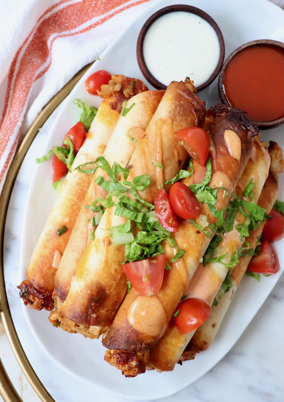 cooked taquitos stacked up on a plate topped with buffalo sauce, tomatoes, and shredded lettuce
