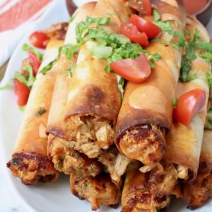 cooked buffalo chicken taquitos stacked up on a plate topped with cilantro and diced tomatoes