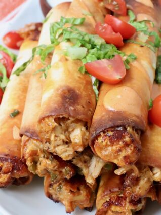 cooked buffalo chicken taquitos stacked up on a plate topped with cilantro and diced tomatoes