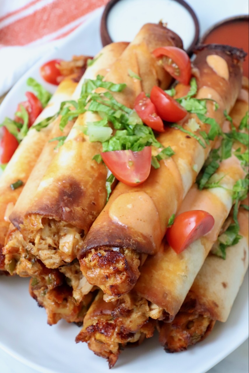 air fried chicken taquitos stacked up on a plate topped with chopped lettuce and tomatoes