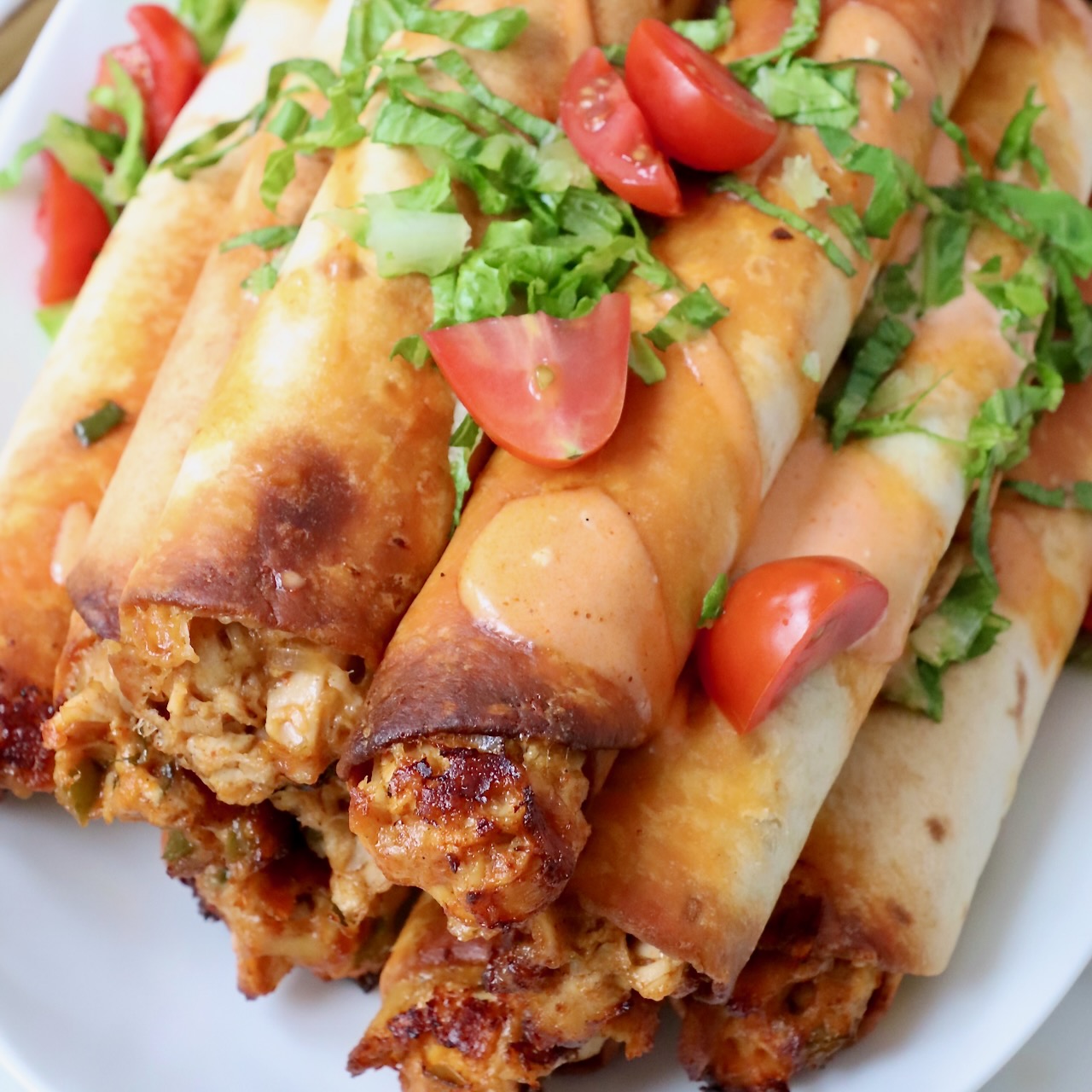 buffalo chicken taquitos stacked up on a plate topped with shredded lettuce and diced tomatoes