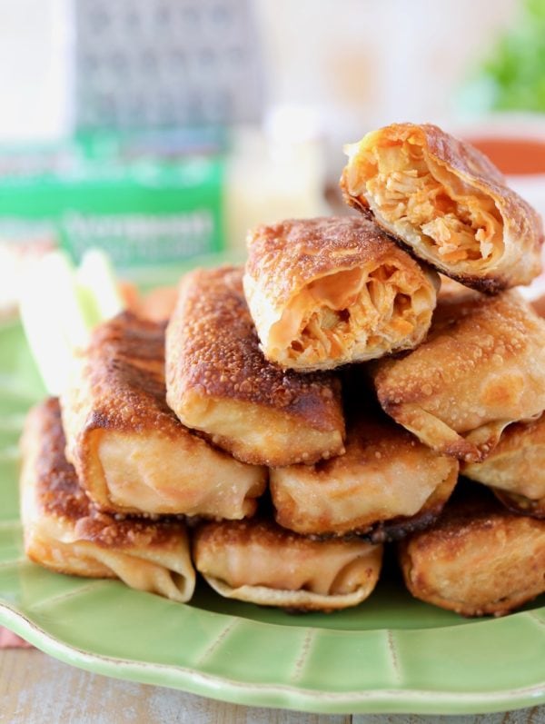 Buffalo chicken egg rolls piled up on a green plate with one egg roll cut in half on top, with cheddar cheese and a cheese grater in the background